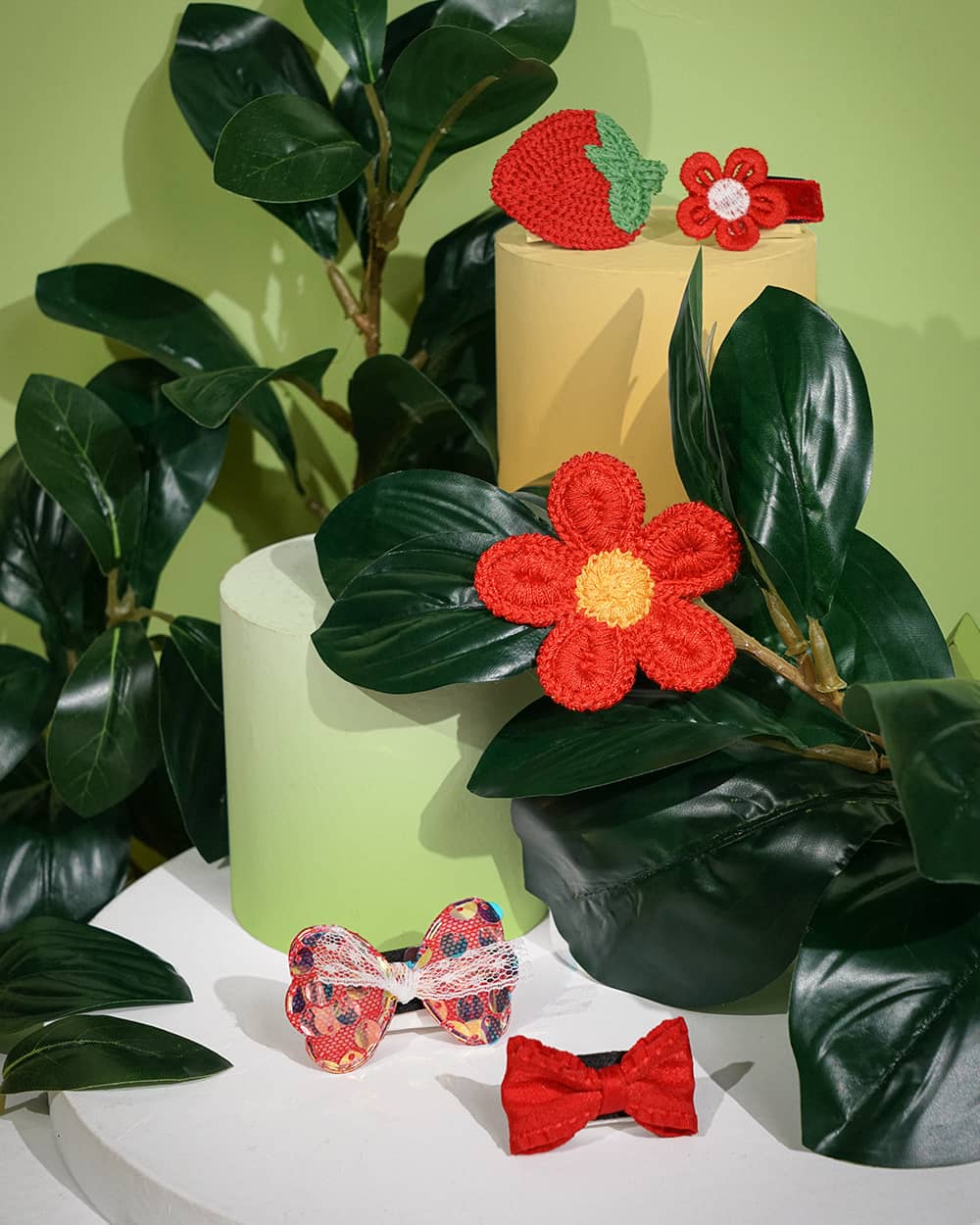 a set of vibrant pet hair accessories, including crocheted red flowers, a patterned bow, and a solid red bow. The accessories are displayed against a backdrop of lush, green leaves, creating a lively and nature-inspired setting. The hair clips are made of high-quality materials and are designed to add a touch of playful style to any pet's look.